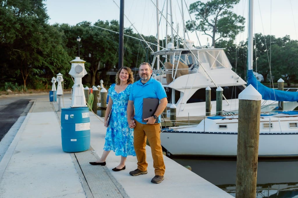 Anna and Bryan Cirlot holding hands on a Gulf Coast marina dock, each with a laptop