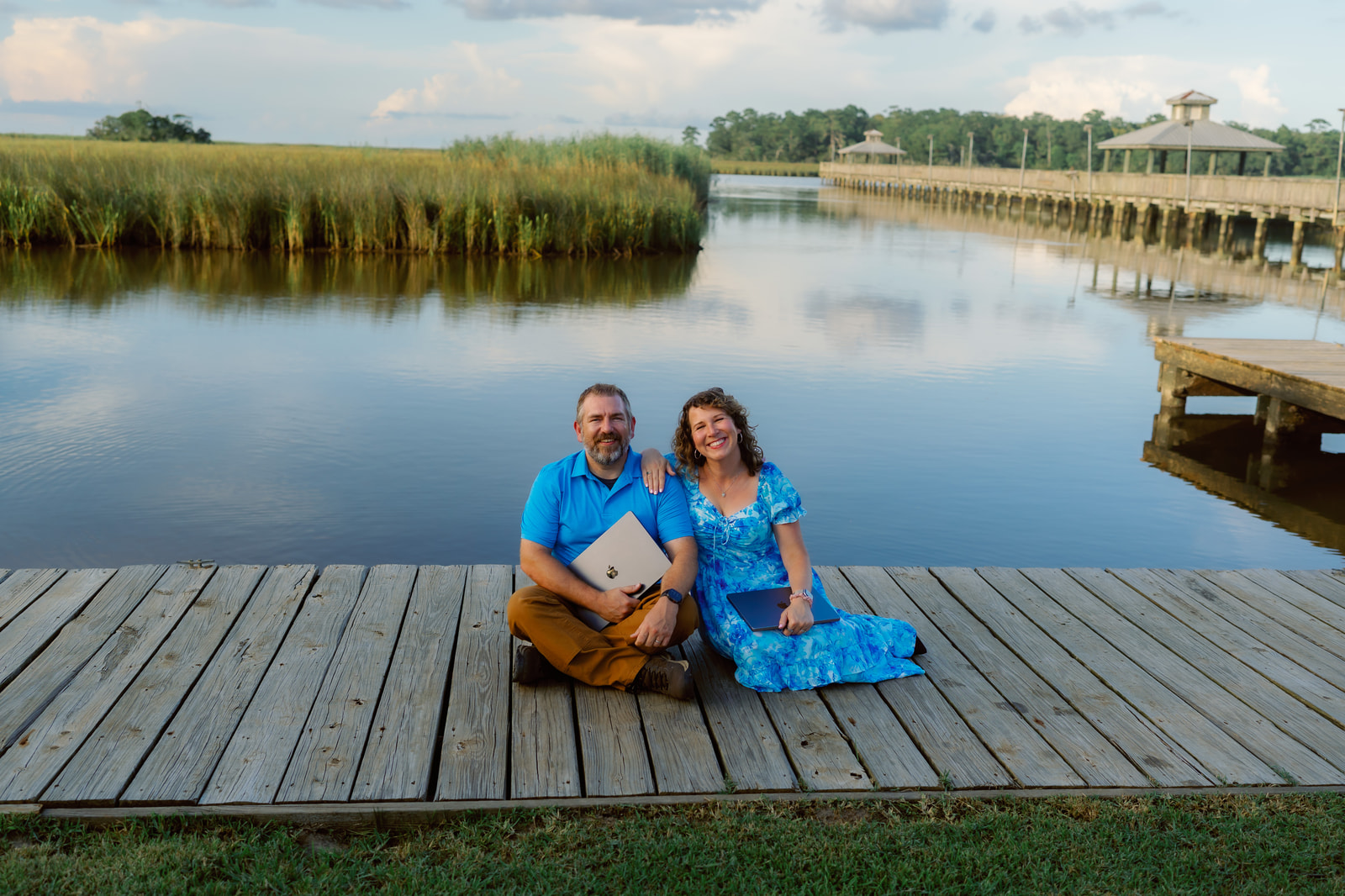 Anna and Bryan Cirlot sitting on a Gulf Coast dock with laptops, marsh and pier behind them